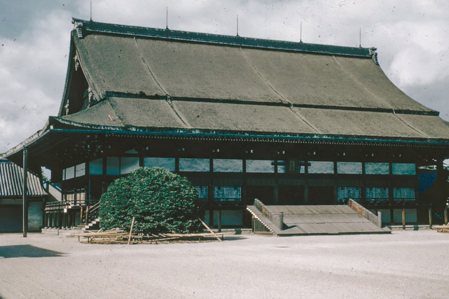 Traditionell japansk byggnad med stort vasstak och trätrappa, omgiven av grus och buskage.